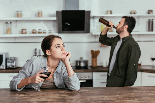 Выборочный фокус задумчивой женщины с бокалом вина и мужем, пьющим пиво на кухне
