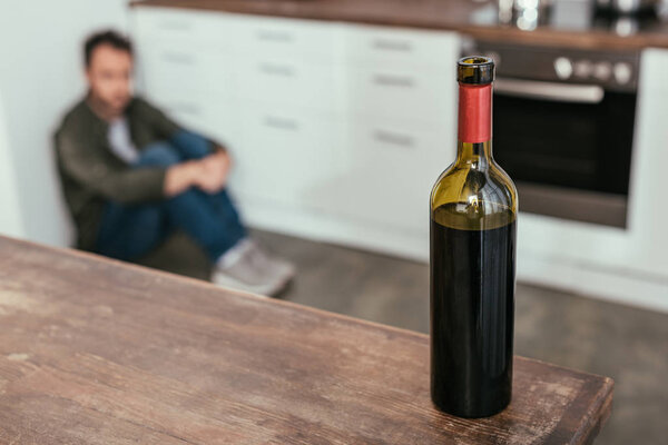Selective focus of wine bottle on table and man sitting on kitchen floor