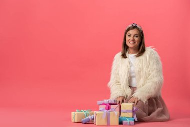 Çekici ve gülümseyen kadın pembe arka planda hediyelerin yanında oturuyor. 