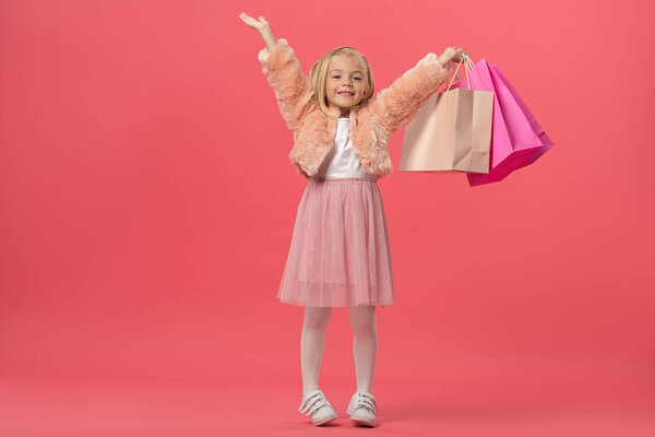 милый и улыбающийся ребенок с протянутыми руками, держащий сумки для покупок на розовом фоне
 