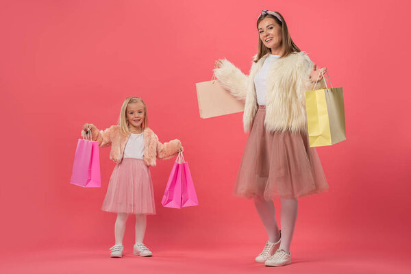 smiling daughter and mother holding shopping bags on pink background 