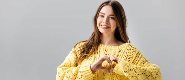 panoramic shot of smiling girl in yellow sweater showing heart sign isolated on grey clipart