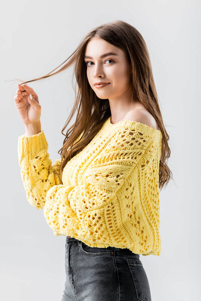 attractive, flirty girl looking at camera while touching hair isolated on grey