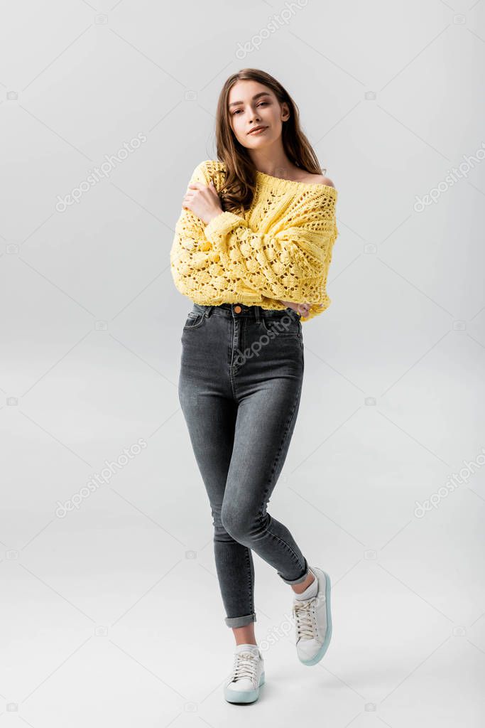 full length view of confident young woman looking up while hugging herself isolated on grey