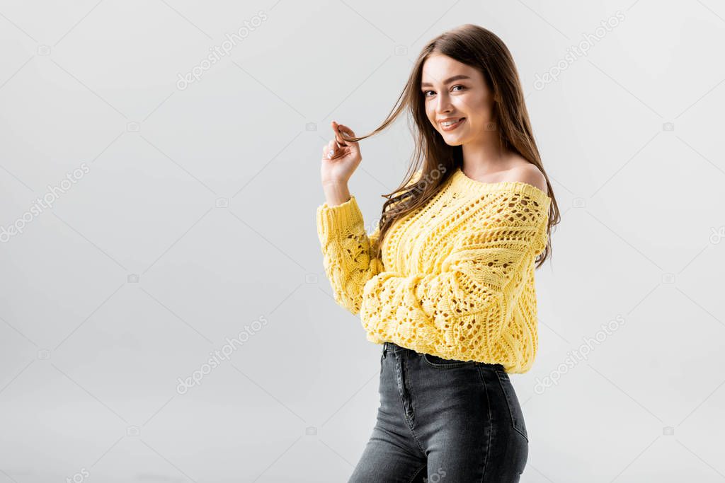 attractive, flirty girl looking at camera while touching hair isolated on grey