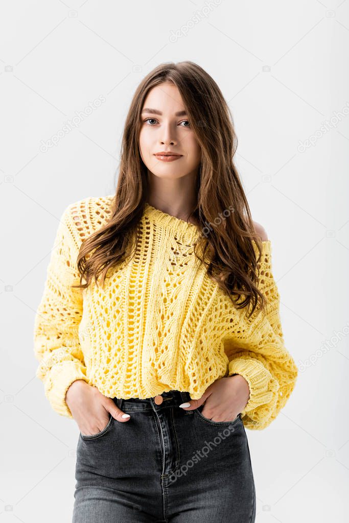 positive girl in yellow sweater standing with hands in pockets and looking at camera isolated on grey