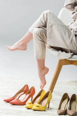 Çıplak ayaklı bir kızın, gri arka plandaki ayakkabı koleksiyonunun yanındaki sandalyede oturması.