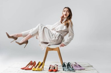Gri arka plandaki ayakkabı koleksiyonunun yanında yükseltilmiş bacaklarla sandalyeye oturmuş şaşırmış bir kız.