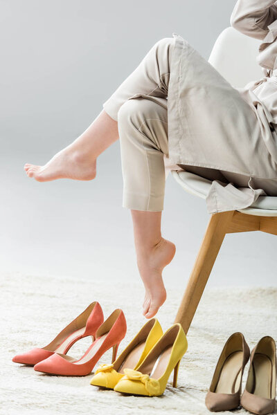 cropped view of barefoot girl sitting on chair near collection of shoes on grey background