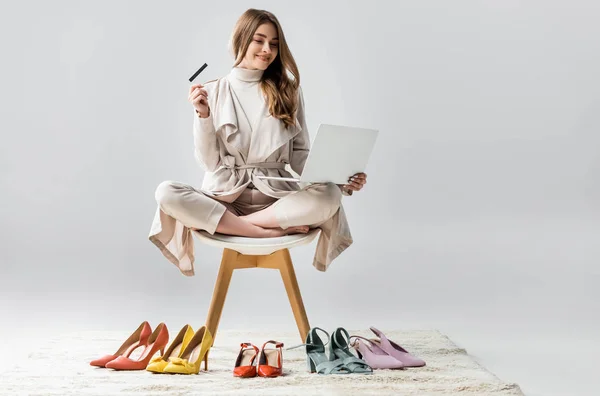 陽気な女の子は蓮のポーズで椅子に座ってノートパソコンとクレジットカードを持ってグレーの背景に靴のコレクションの近くに — ストック写真