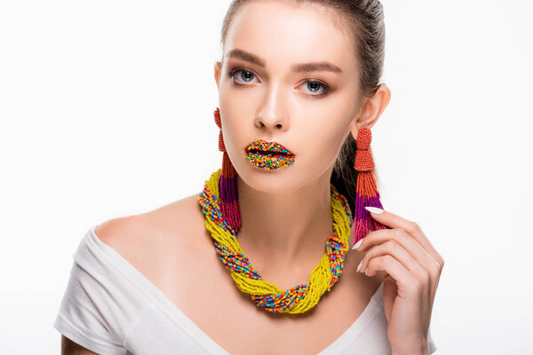 beautiful girl in beaded accessories, with beads on lips, touching earring and looking at camera isolated on white