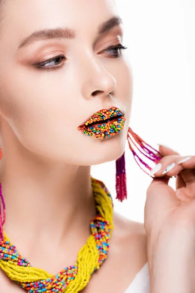 beautiful girl in beaded accessories, with beads on lips, touching earring and looking  away isolated on white