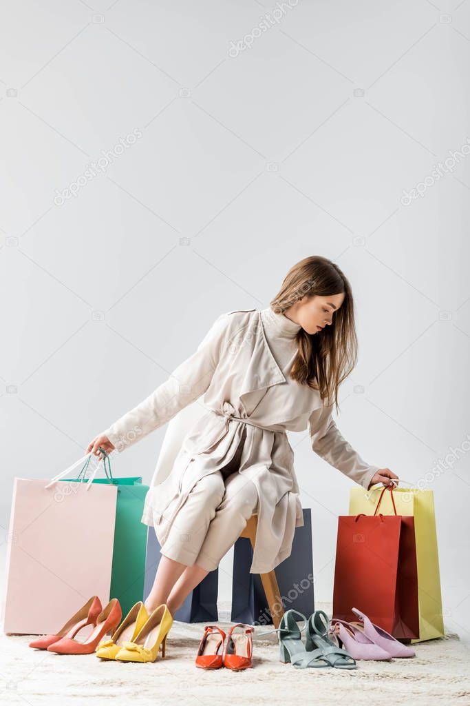 trendy young woman holding shopping bags while sitting near collection of shoes on grey background