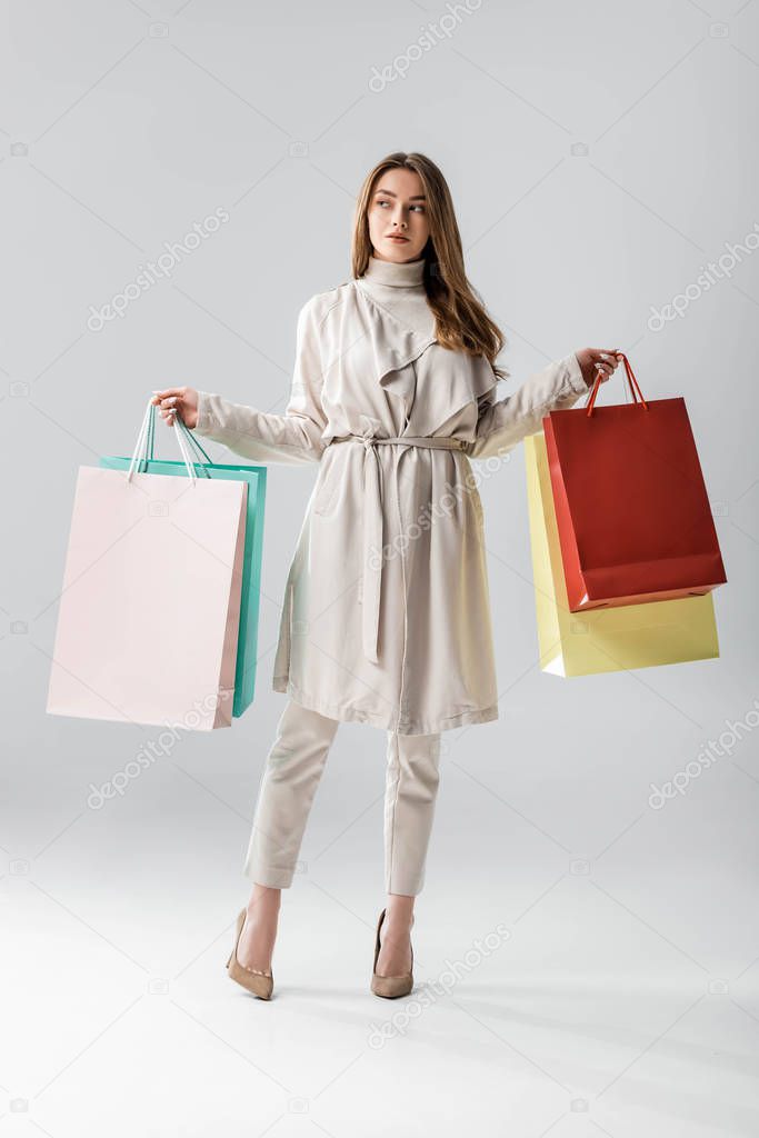 full length view of stylish girl looking away while holding shopping bags on grey background