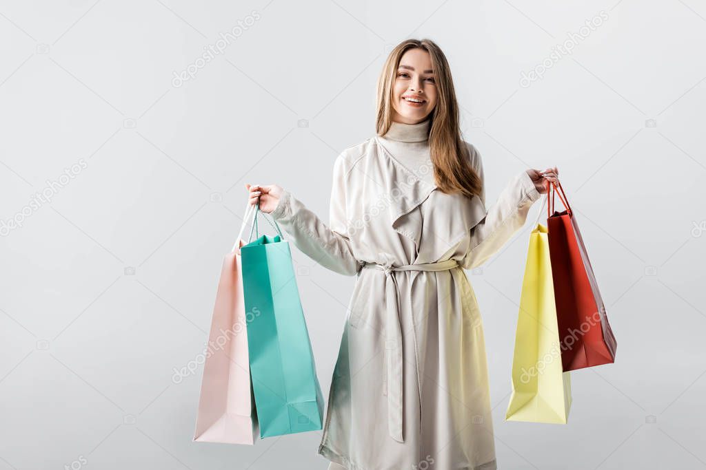 cheerful, stylish girl looking at camera while holding shopping bags isolated on grey