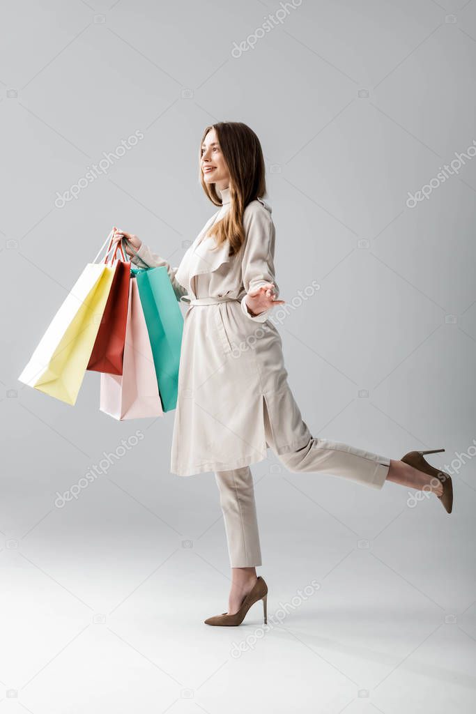 full length view of happy girl in stylish cape holding shopping bags on grey background