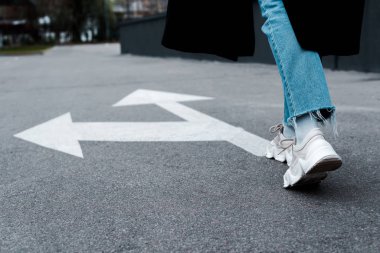 cropped view of woman in jeans walking near directional arrows on asphalt  clipart