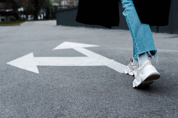cropped view of woman in jeans walking near directional arrows on asphalt 