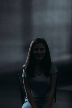 Genç bir kadın karanlık arka planda otururken kameraya gülümsüyor.