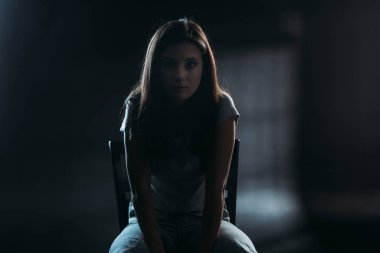 Siyah arka planda, karanlıkta bir sandalyede oturmuş kameraya bakan umutsuz genç bir kadın.