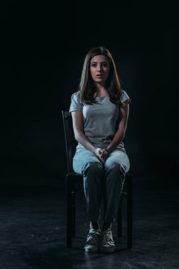 Üzgün genç bir kadın, siyah arka planda, karanlıkta bir sandalyede otururken kameraya bakıyor.