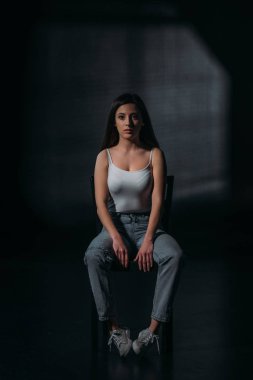 Depresyonda olan kadın, siyah arka planda, karanlıkta bir sandalyede otururken kameraya bakıyor.