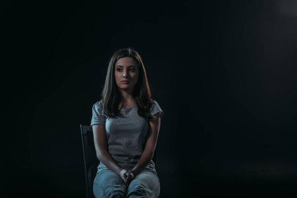 despaired woman looking away while sitting on chair in darkness isolated on black