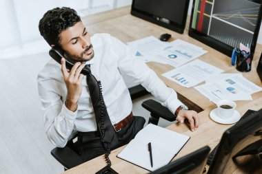 Telefonda konuşan ve bilgisayarın yanında grafiklerle oturan iki ırklı tüccarın yüksek açılı görüntüsü 