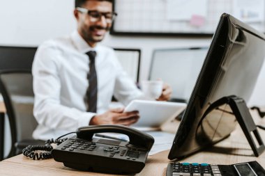 Telefon ve hesap makinesinin masadaki seçici odağı ve arka planda dijital tableti olan gülümseyen tüccar 