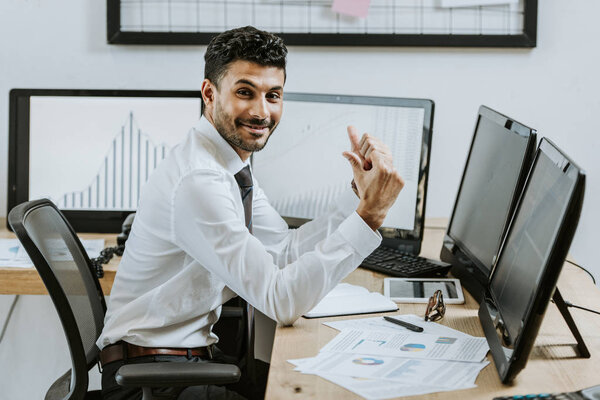 smiling bi-racial trader showing thumbs up and sitting near computers with graphs 