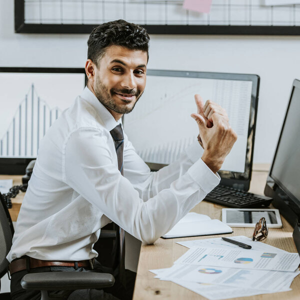 smiling bi-racial trader showing thumbs up and sitting near computers with graphs illustration 
