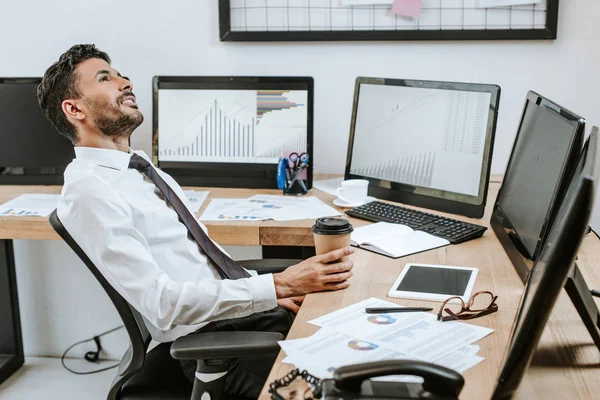 Sonriente Racial Comerciante Sosteniendo Taza Papel Sentado Cerca Computadoras Con — Foto de Stock