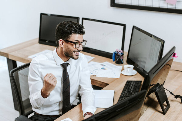 high angle view of smiling bi-racial trader showing yes gesture and looking at computer 