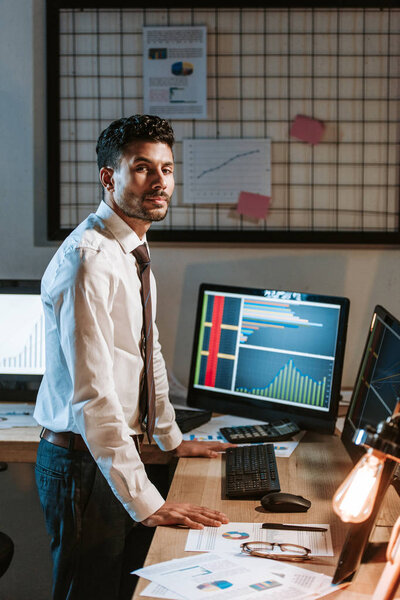 bi-racial trader standing near computers with graphs and looking at camera 
