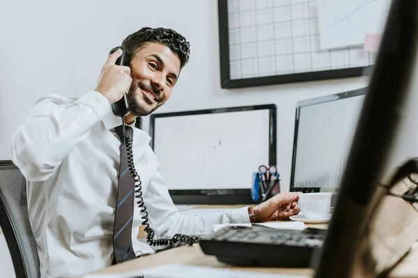 Uśmiechnięty Dwurasowy Przedsiębiorca Rozmawiający Przez Telefon Siedzący Przy Stole — Zdjęcie stockowe