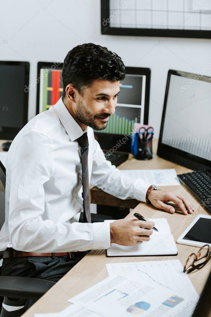 smiling bi-racial trader writing and sitting near computers with graphs 