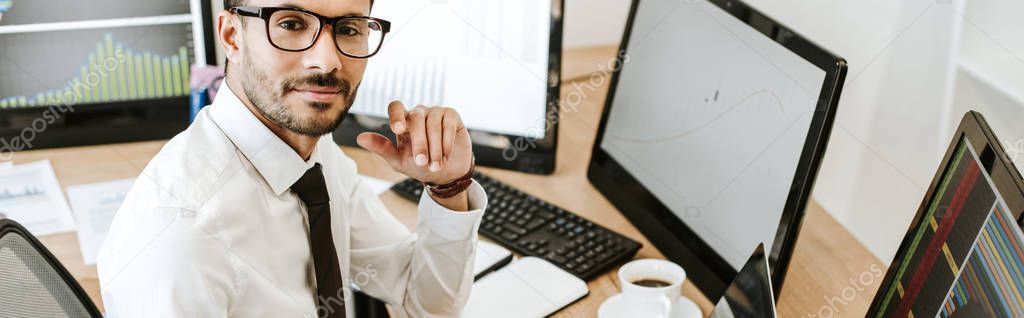 panoramic shot of smiling bi-racial trader sitting near computers and looking at camera 