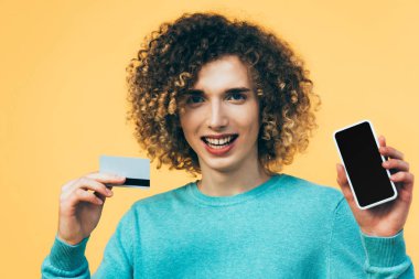 Sırıtkan kıvırcık ergen elinde akıllı telefon ve kredi kartıyla sarıya izole edilmiş.