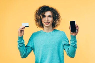 Sırıtkan kıvırcık ergen elinde akıllı telefon ve kredi kartıyla sarıya izole edilmiş.
