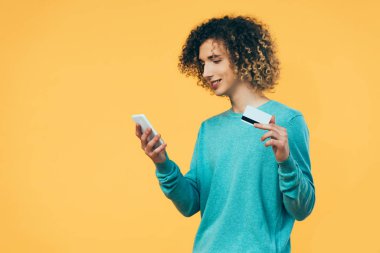 Sırıtkan kıvırcık ergen elinde akıllı telefon ve kredi kartıyla sarıya izole edilmiş.