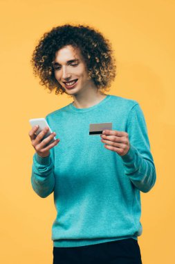 Sırıtkan kıvırcık ergen elinde akıllı telefon ve kredi kartıyla sarıya izole edilmiş.