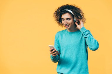 Gülümseyen kıvırcık ergen akıllı telefon kullanıyor ve sarı kulaklıkla müzik dinliyor.