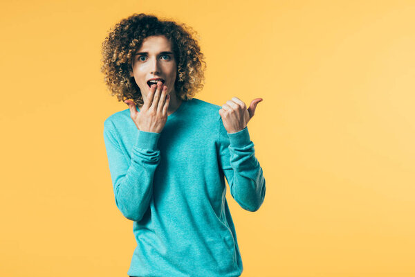 shocked curly teenager covering mouth with hand and pointing with finger aside isolated on yellow