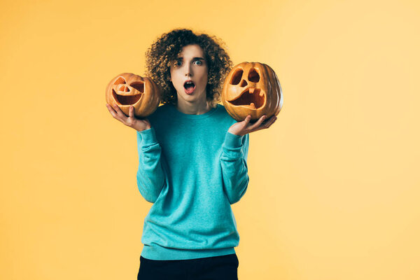 shocked curly teenager holding Halloween pumpkins isolated on yellow