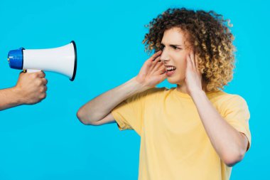 cropped view of man holding loudspeaker near curly teenager covering ears isolated on blue clipart