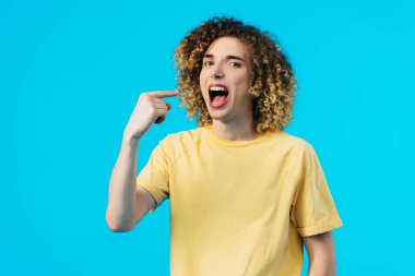 hungry curly teenager pointing with finger at mouth isolated on blue clipart