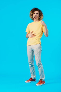 hungry curly teenager pointing with finger at mouth and touching stomach isolated on blue clipart