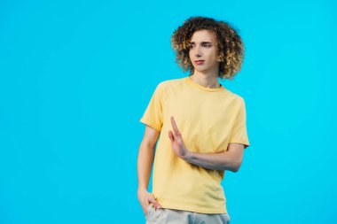 confused curly teenager with hand in pocket showing stop gesture isolated on blue