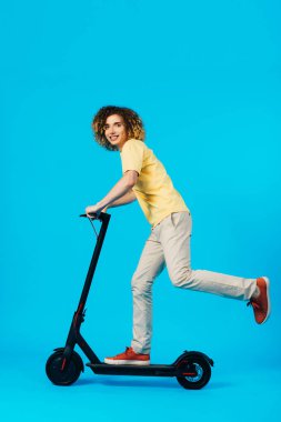 Mavi arka planda elektrikli scooter süren mutlu kıvırcık gencin yan görüntüsü