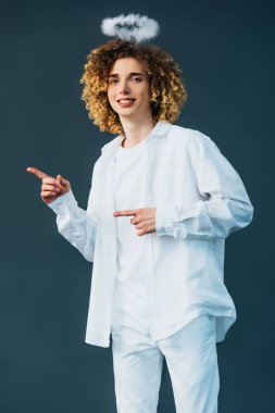 smiling curly teenager in angel costume with halo above head pointing with fingers isolated on green clipart